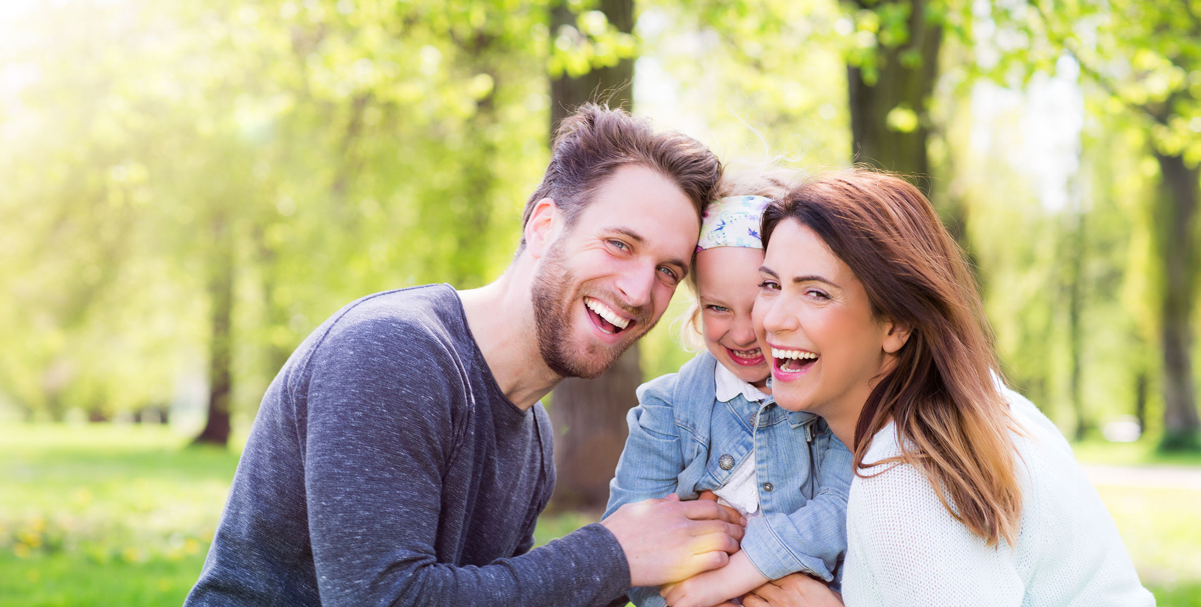 A family of four in a park, with the father holding the child and smiling at the camera.