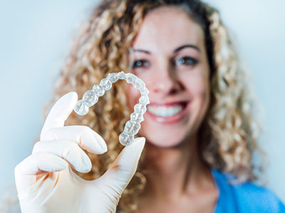 A woman, smiling and wearing a blue surgical mask, holds up an open package containing a clear dental retainer.