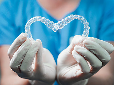 The image shows a pair of clear dental braces held by someone wearing white gloves and a blue shirt, with a heart shape formed by the person s hands.
