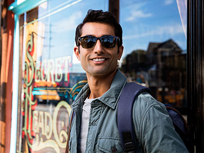 The image features a man wearing sunglasses and a backpack, standing in front of a storefront with a sign that reads Secret Garden. He is posing for the photograph.